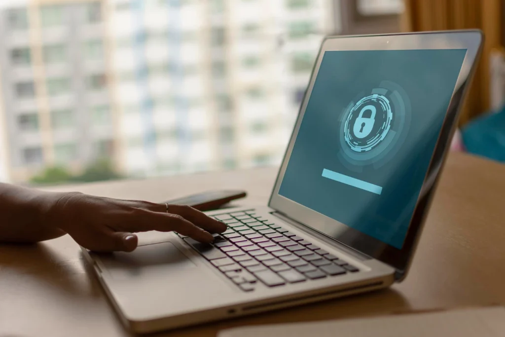 Kéz laptop billentyűzetén, a képernyőn biztonsági zár ikon látható a hozzáférés védelmét jelképezve.